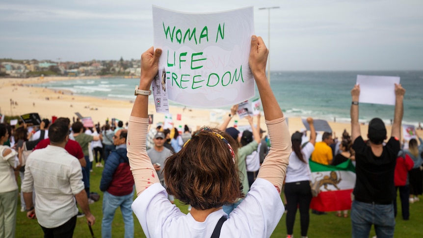 A woman in a white shirt holds a sign that reads "Woman Life Freedom" at a protest held on Bondi Beach.