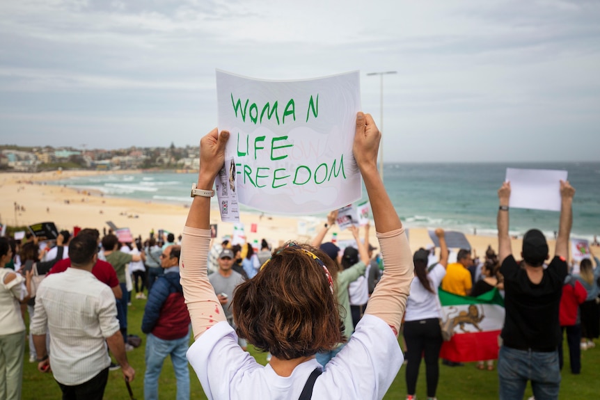 A woman in a white shirt holds a sign that reads "Woman Life Freedom" at a protest held on Bondi Beach.