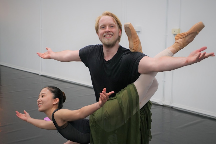 A blonde ballet dancer smiling with his arms out supporting a ballerina on his legs.