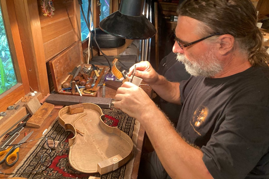 A man using a small clamp to hold two pieces of a violin together at his workshop bench.