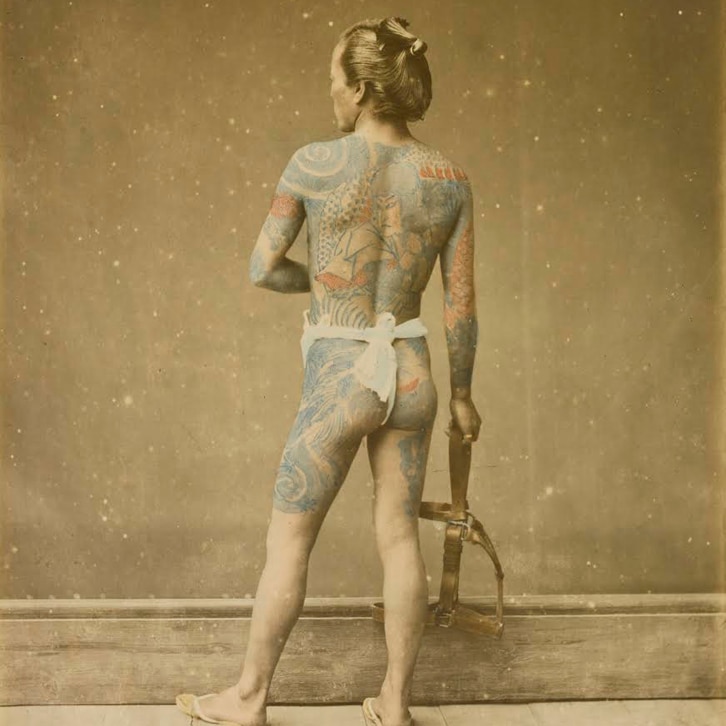 A Japanese man wearing only a loincloth stands with his back to the camera, showing off izerumi tattoos.