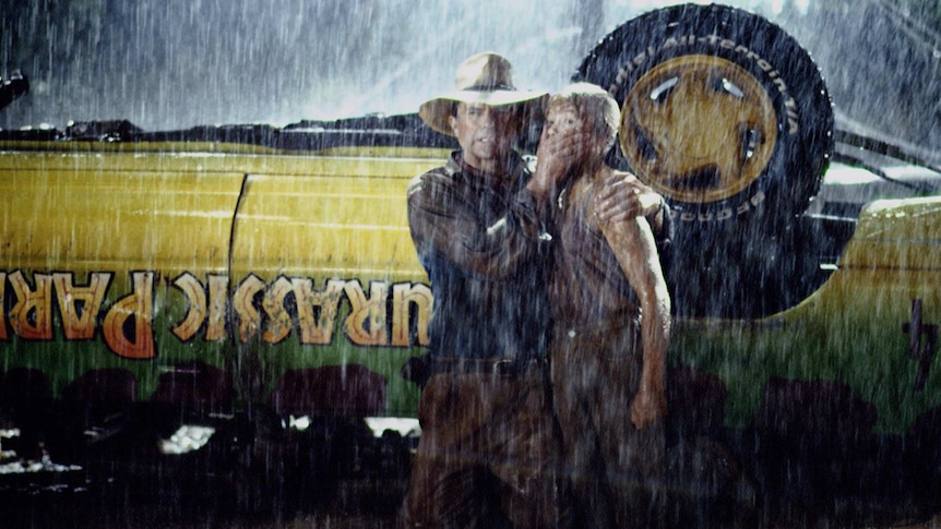 A man in a ranger's hat stifling the scream of a young girl, in front of an overturned vehicle with the 'Jurassic Park' logo.