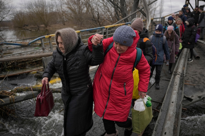 Women hold hands while crossing the Irpin river on an improvised path under a bridge