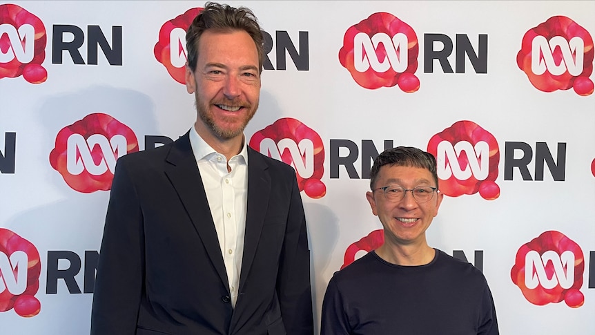 A tall man in a suit with brown hair and a beard stands next to a shorter man in a black shirt and glasses, both smile 