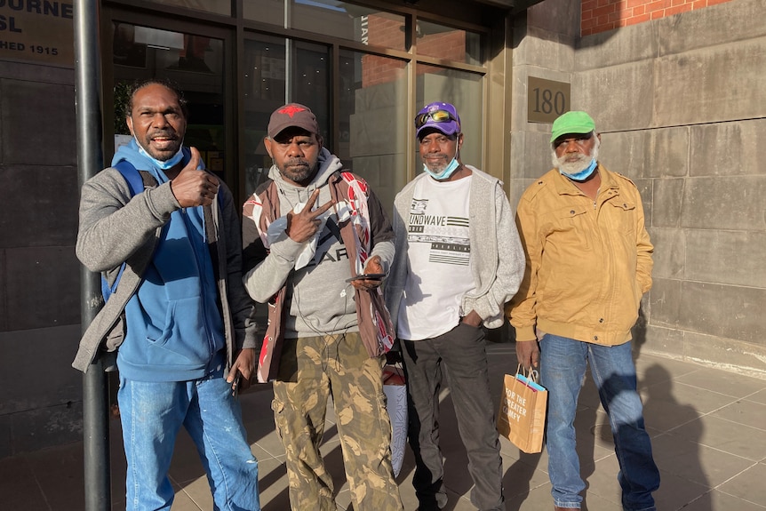 Four Arnhem Land men smile, two with thumbs up, face masks pulled down, stand outside building.