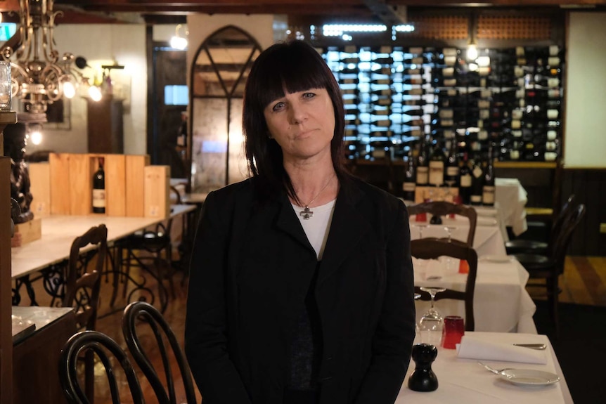 A woman with dark hair sits at a restaurant table with a mirror behind her.