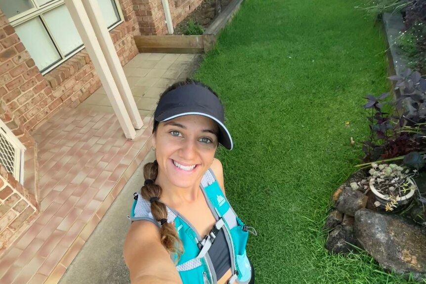 A selfie of a young woman smiling up at the camera, wearing a dark cap and aqua blue training vest. 