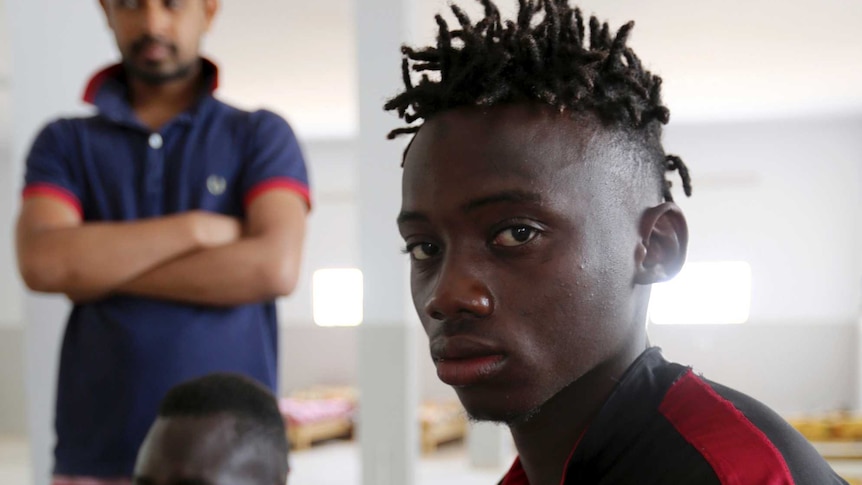 A Malian survivor of a shipwreck stares at the camera as two other men sit behind him.