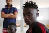 A Malian survivor of a shipwreck stares at the camera as two other men sit behind him.