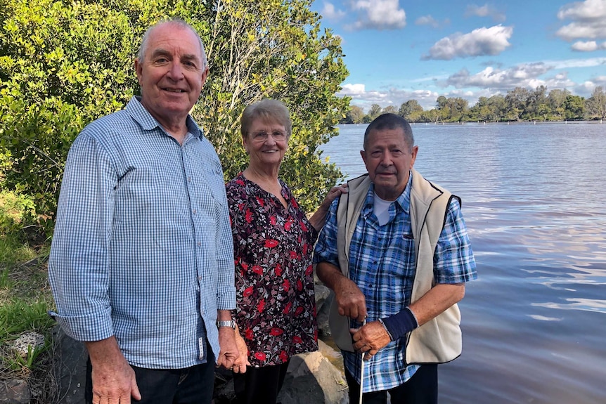 Two older men and a woman stand on the banks of a river with a tree behind them, all smiling.