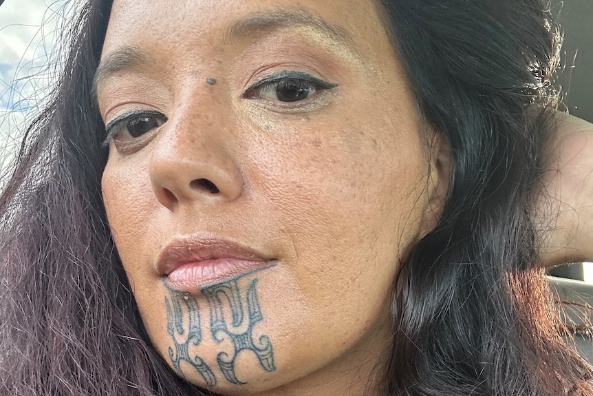 A woman with long dark hair and a tattoo on her chin looks at the camera.