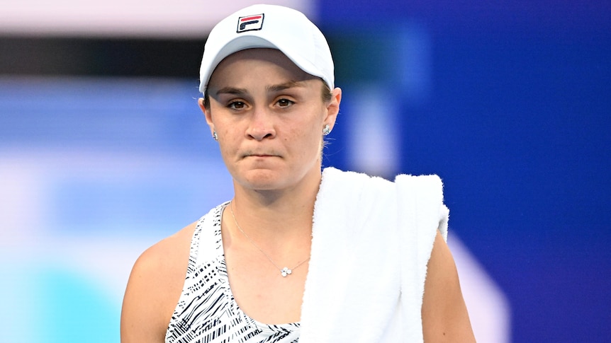 An Australian female tennis player walks along a court with a towel over her shoulder. 