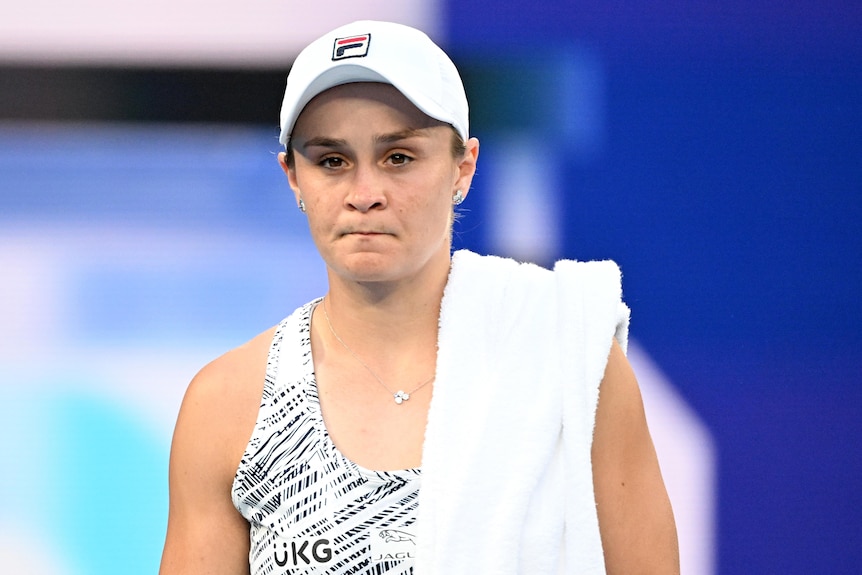 An Australian female tennis player walks along a court with a towel over her shoulder. 