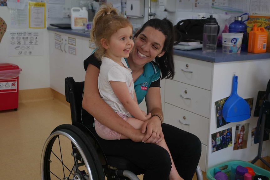 A young woman in a wheelchair cuddles a young girl at an early childhood centre.