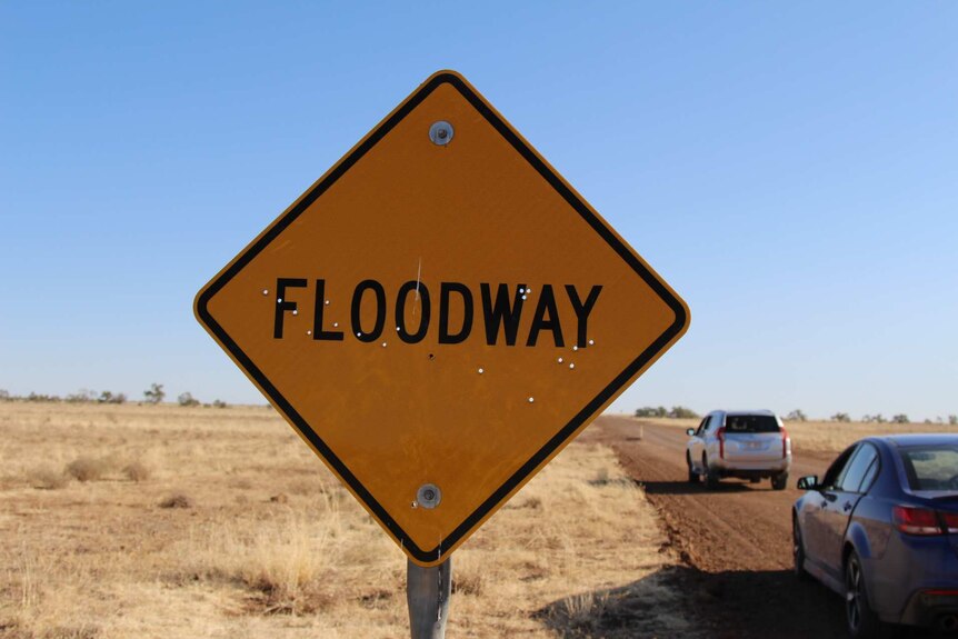 A sign reading "floodway" in front of a dusty road and flat field of dry grass.