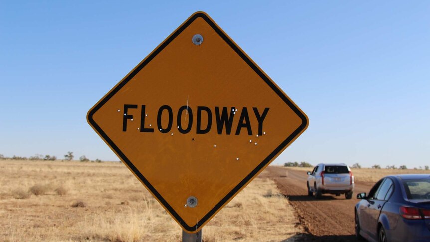 A sign reading "floodway" in front of a dusty road and flat field of dry grass.