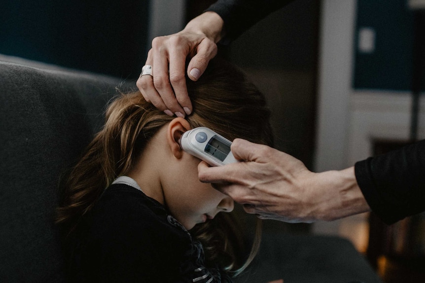 Close up of an adult's hand holding an in-ear thermometer to a child's ear.