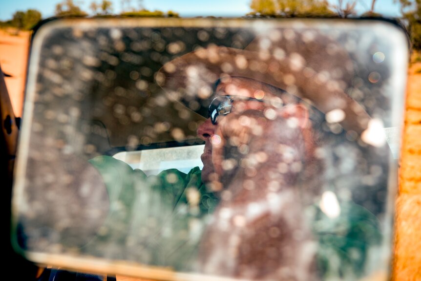 Boyd Webb is seen driving, reflected in a side mirror covered in flecks of mud and dust.