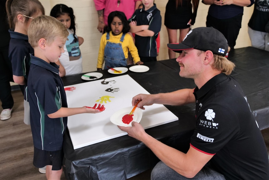 A man in a cap and a dark, branded polo shirt helps a youngster do a spot of finger painting.