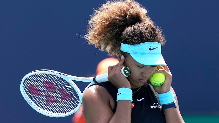 A visor-wearing tennis player covers her ears with her hands as she also holds her racquet and a tennis ball.