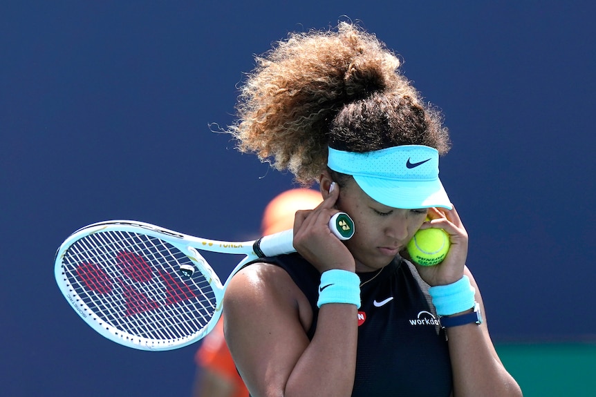 Una jugadora de tenis con visera se cubre las orejas con las manos mientras sostiene su raqueta y una pelota de tenis.