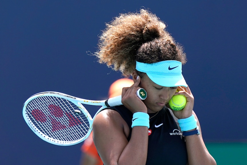 A visor-wearing tennis player covers her ears with her hands as she also holds her racquet and a tennis ball.