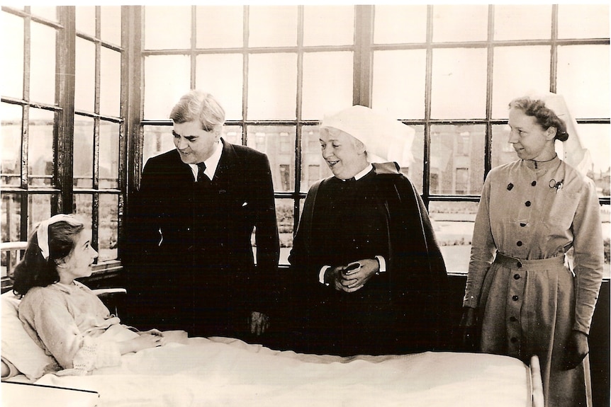 Black and white photo of a man in a suit and two women wearing nurses' hats standing at the bedside of a young woman.