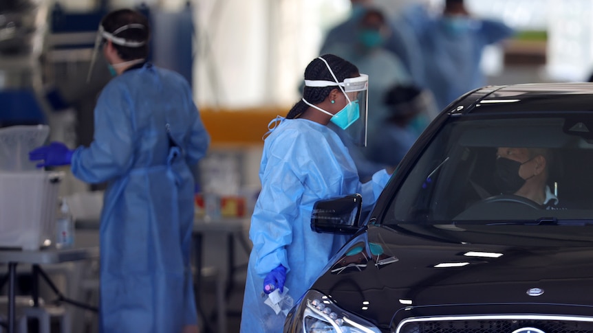 A female COVID tester holds a swab up to a car window. She is wearing PPE, a mask and a face shield