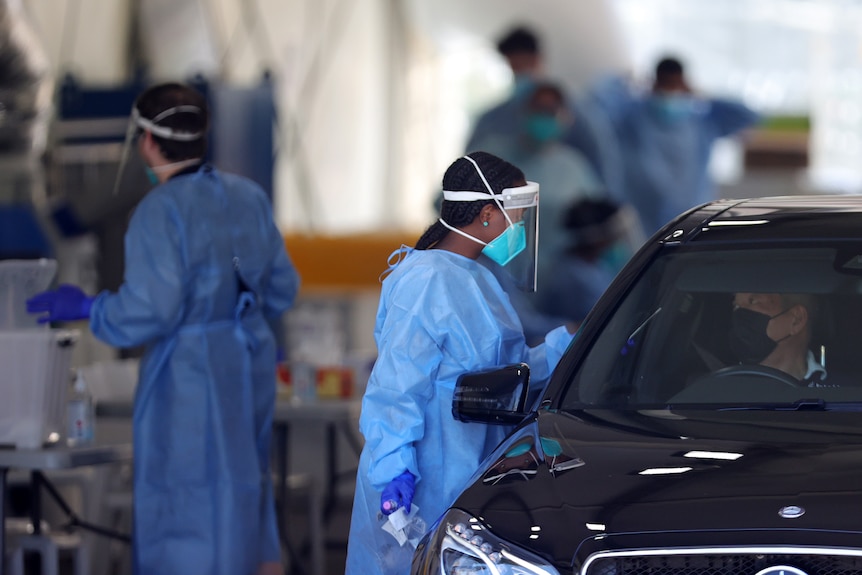 A female COVID tester holds a swab up to a car window. She is wearing PPE, a mask and a face shield