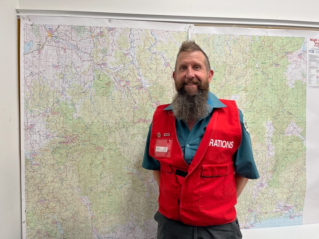 Um homem de cabelos castanhos e barba longa, vestindo um colete de controlador de operações, está em frente a um mapa.
