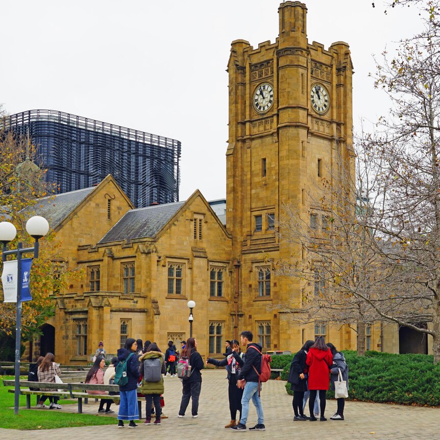 About a dozen student are seen talking in a courtyard, in front of a tall sandstone clock tower at the University of Melbourne.