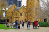 About a dozen student are seen talking in a courtyard, in front of a tall sandstone clock tower at the University of Melbourne.