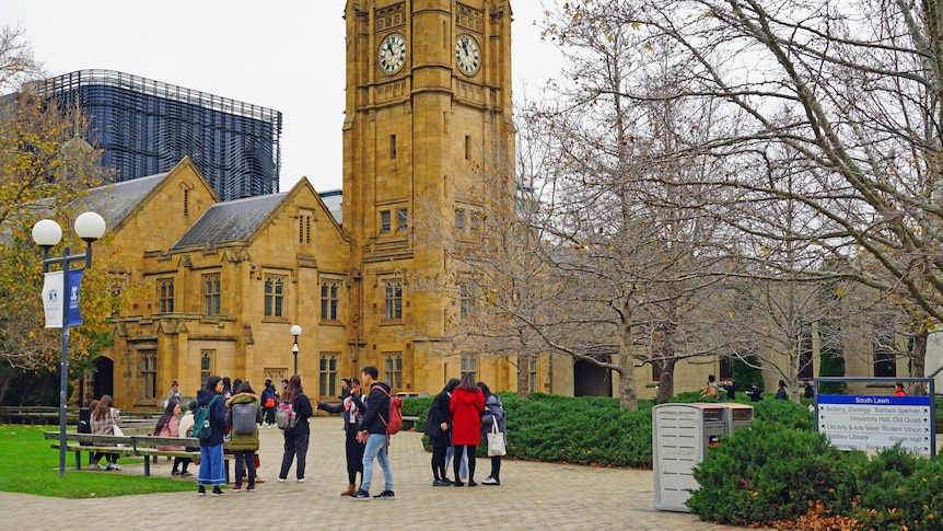 About a dozen student are seen talking in a courtyard, in front of a tall sandstone clock tower at the University of Melbourne.