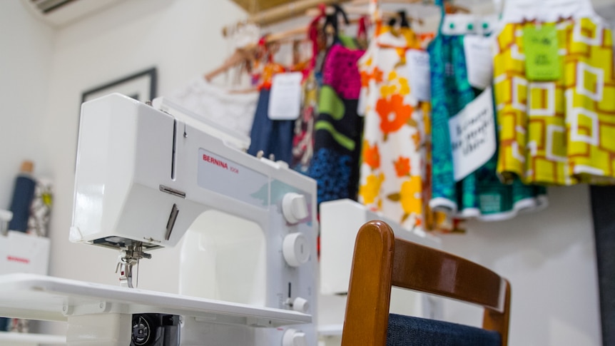 Sewing machine sits on a workbench in front of dresses.