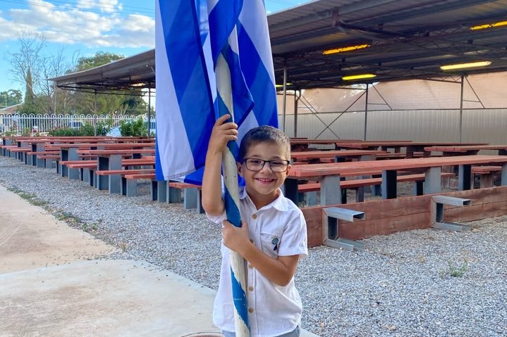 A child wearing a white shirt and black glasses holds a Greek flag, smiling. 