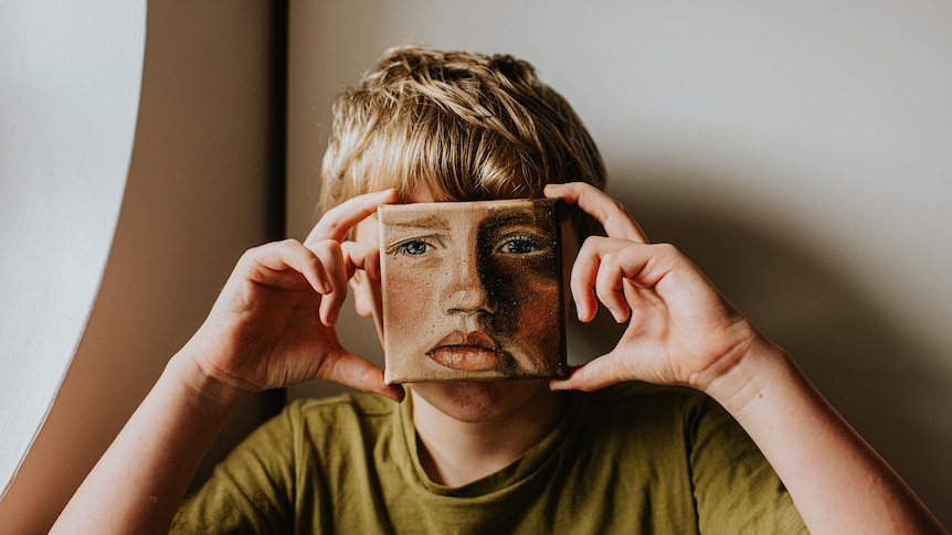 Little boy covers his face with a painting. The painting is a depiction of his face with a frown on it.