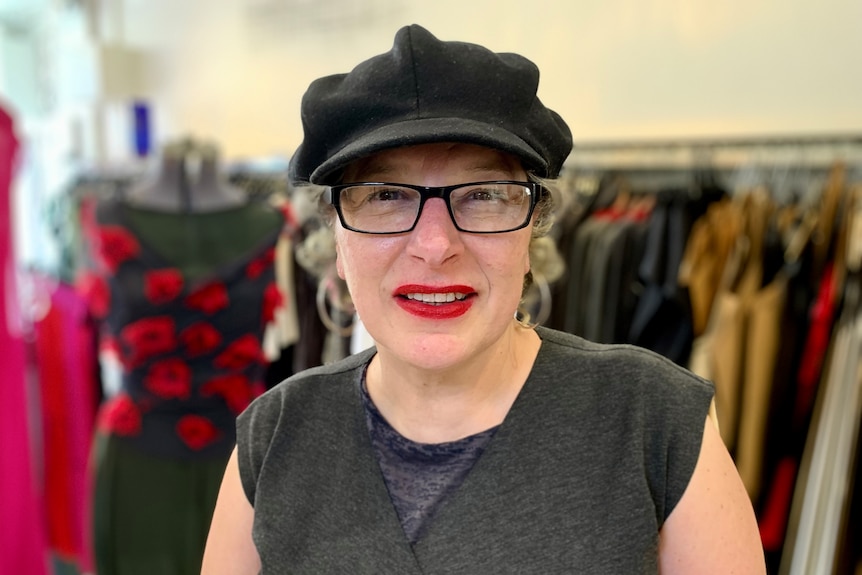 Viv wears a little black cap and looks intently in the camera, in front of a rack of clothing