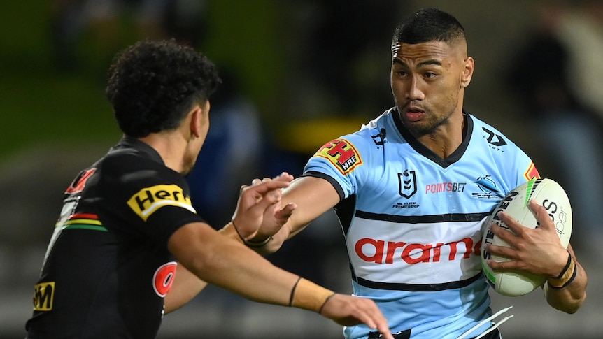A Cronulla NRL player holds the ball as he attempts to fend off a Penrith opponent.