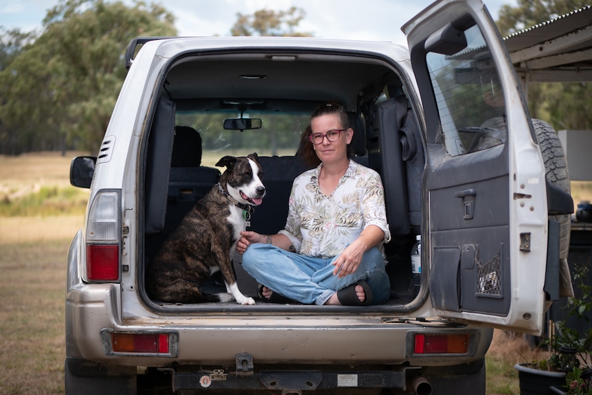 A woman sits in the back of a four wheel drive with a dog. 