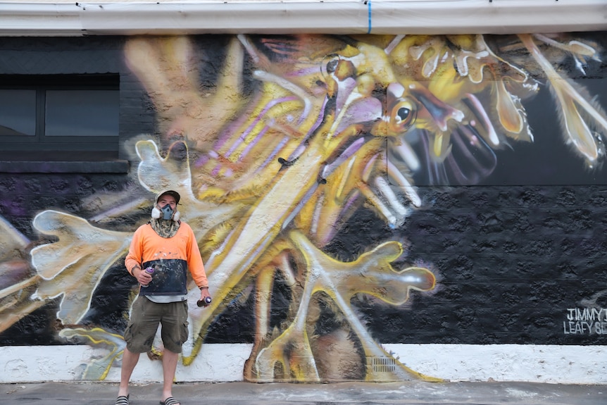 Man in high vis orange t-shirt and hat standing in front of his spray-painted mural of leafy sea dragon.