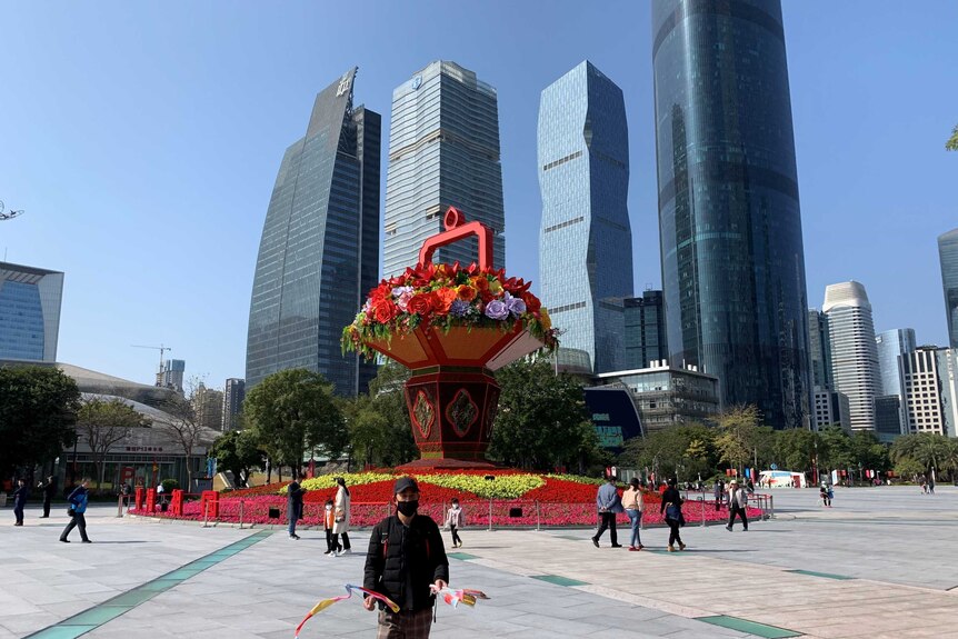 City building in the background in front of a flower-filled square and blue sky.