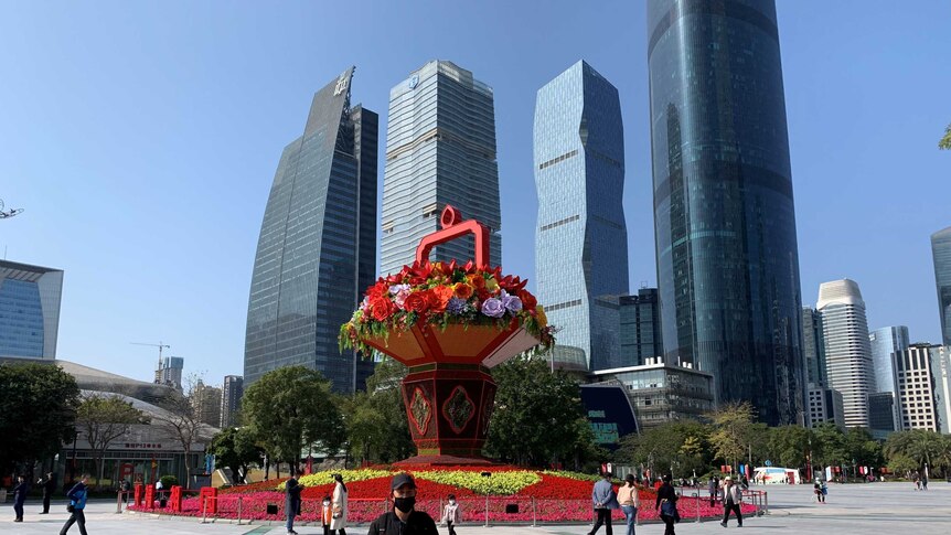 City building in the background in front of a flower-filled square and blue sky.