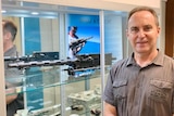 A man in a grey collared shirt, standing in front of a glass case with the gun mounted behind him.