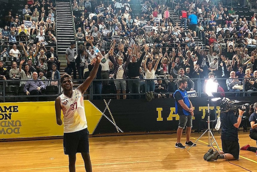 A young man poses with his arm in the air on a basketball court in front of a large cheering crowd.