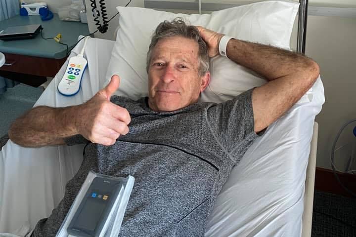 a man with a grey shirt lays in a hospital bed, giving a thumbs up towards the camera