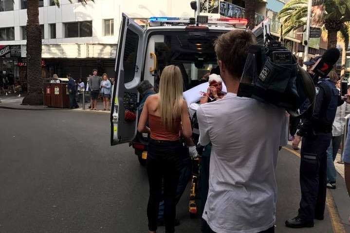 A man with is loaded on a stretcher into the back of an ambulance while a news cameraman films.