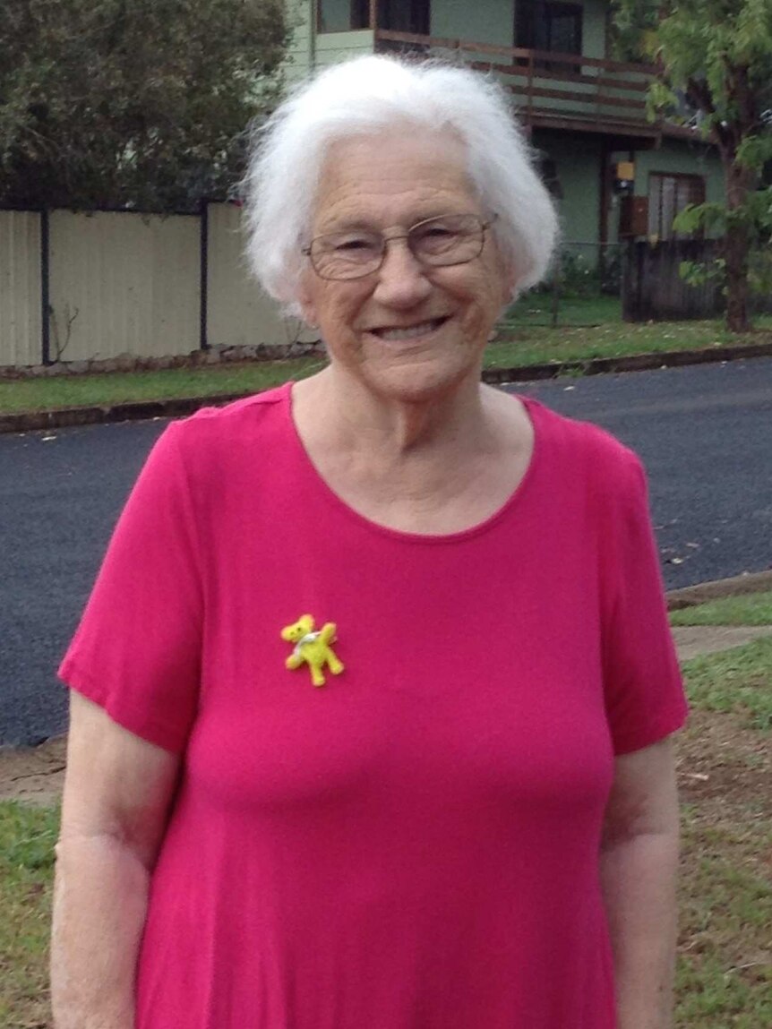 A woman with grey hair and a teddy bear pin on her shirt smiles for the camera in a Queensland street