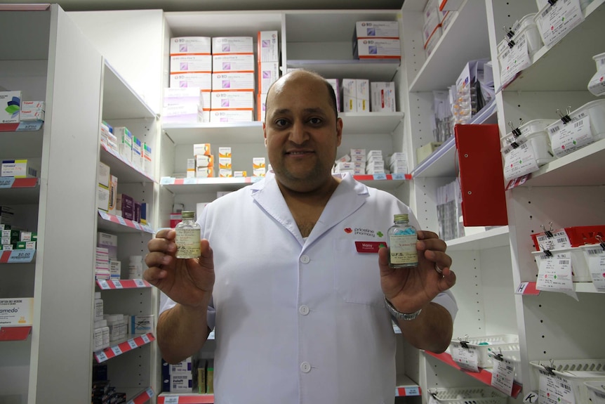 Hany holds the glass bottles and smiles in front of a row of new medication