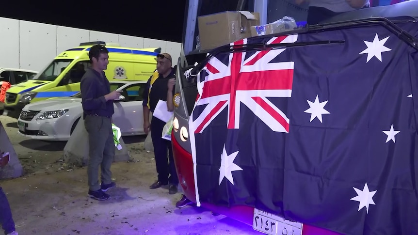 A man with a clipboard talks to another person next to a bus with Australian flag draped on the front