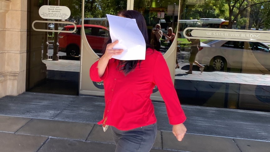 A woman wearing a red top holds a piece of paper in front of her face in front of automatic sliding doors